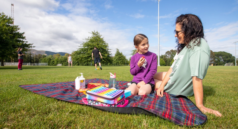 Mother and daughter having a picnic.  People kicking a ball in the background